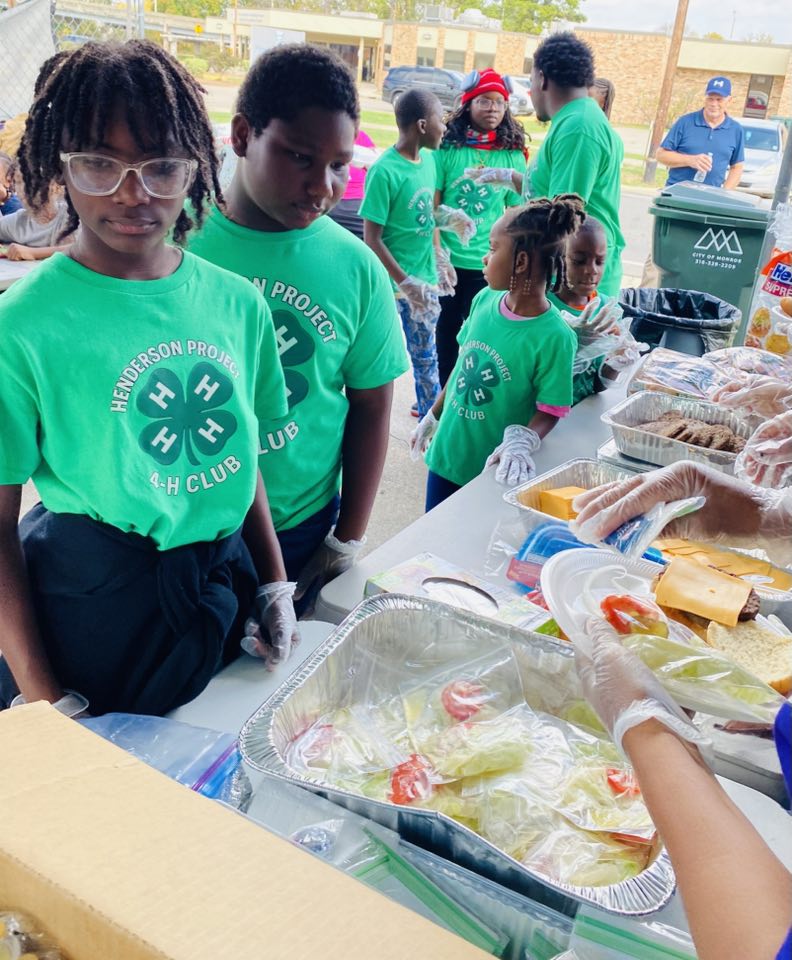 A few kids wear 4-H T-shirts standing in line for food. 4-H Emblem logo.